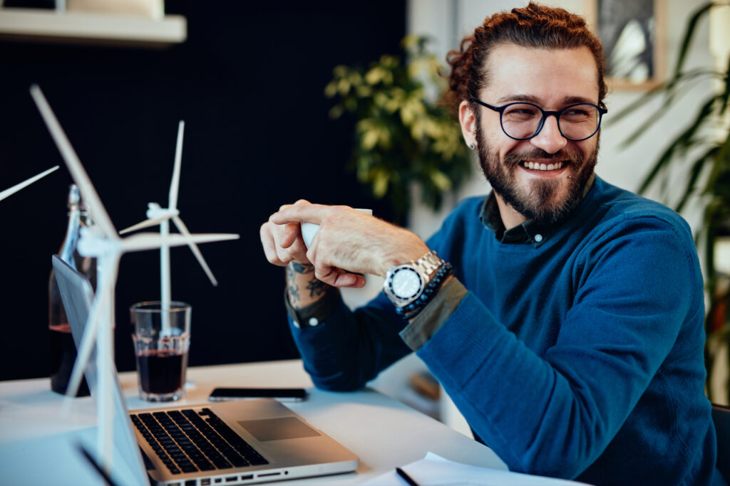 Smiling entrepreneur working at a laptop with wind turbine models and financial templates—accessing free downloadable tools from The William Stanley Group to make smarter business decisions