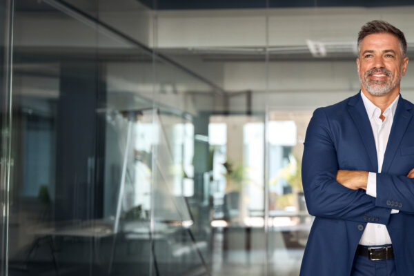Confident business professional standing in a modern glass office, representing The William Stanley Group’s FlexJobs feature on virtual leadership strategies.