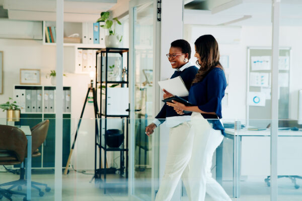 Two professionals smiling and walking through a modern office with glass walls and file shelves, representing The William Stanley Group’s Think Money feature on top financial films.